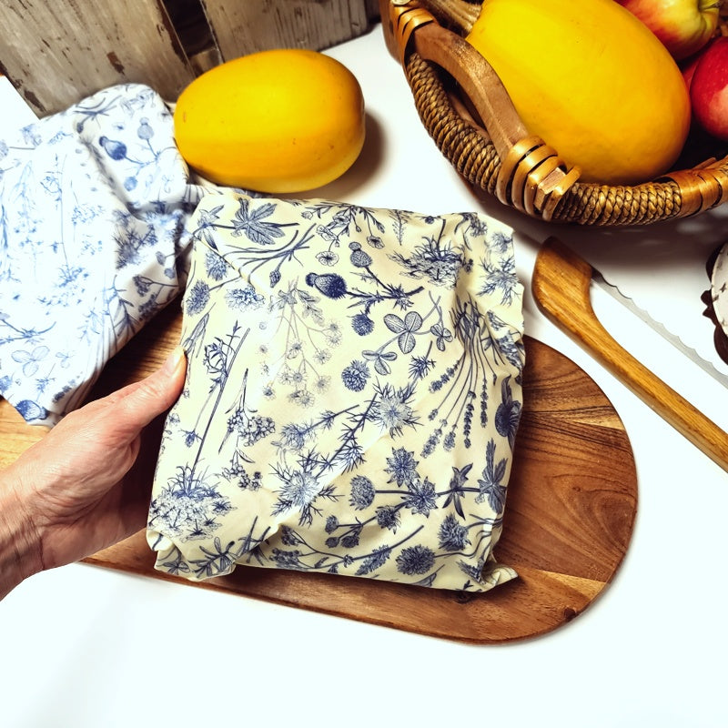 A person holding an extra large beeswax wrap with a blue floral pattern, placed on a wooden board next to some fruits.