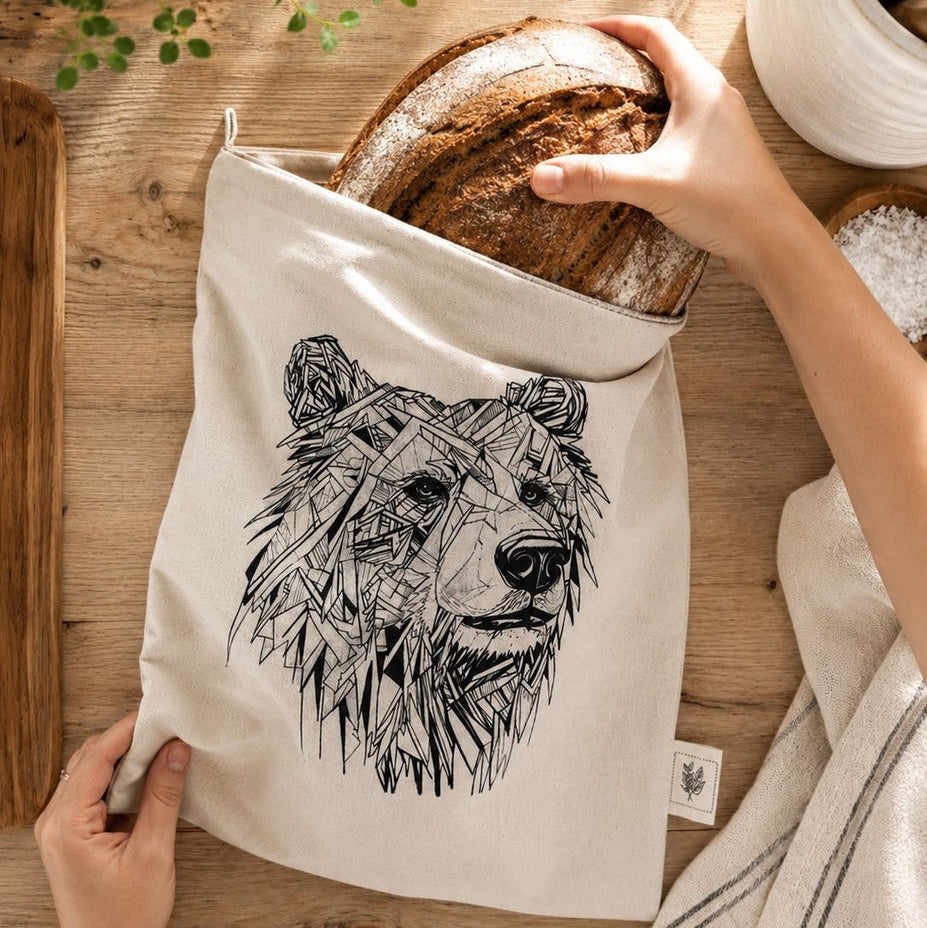A beige canvas bread bag with a black line art bear graphic, being held on a wooden table. A hand is placing a rustic loaf of bread into the top of the bag, and another hand is holding the bottom corner of the bag.