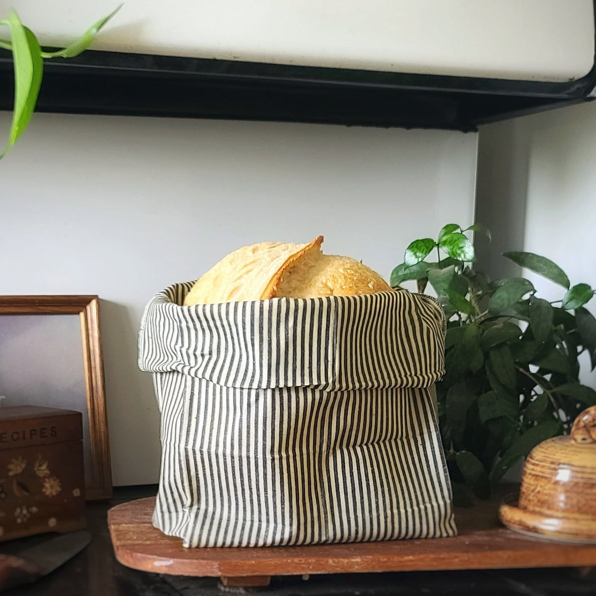 A fabric storage basket with vertical stripes in beige and dark green, filled with tortilla chips, set on a wooden shelf in a home environment with plants and decorative items.